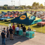 A large pop-up canopy tent with a table set up on an outdoor car show at a grassy field. The canopy is dark teal with yellow diagonal stripes and features the 'American Muscle' logo.