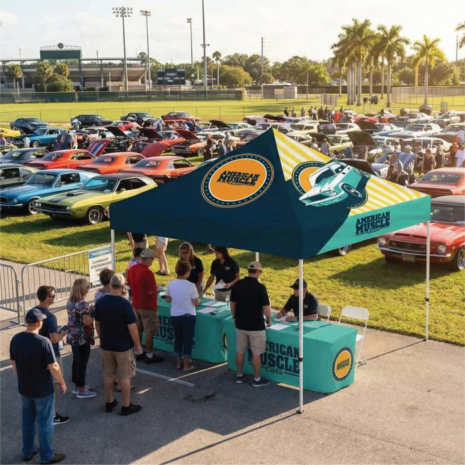 A large pop-up canopy tent with a table set up on an outdoor car show at a grassy field. The canopy is dark teal with yellow diagonal stripes and features the 'American Muscle' logo.