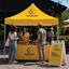 A bright yellow Buzzing Bees outdoor market booth with customers and staff under a branded canopy, with a matching A-frame sign on a paved street in front of a dark building.