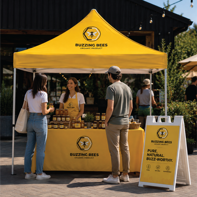 A bright yellow Buzzing Bees outdoor market booth with customers and staff under a branded canopy, with a matching A-frame sign on a paved street in front of a dark building.