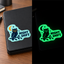 A split-screen product photo showing a black rectangular item with a die-cut sticker that reads “NIGHT SHIFT” and features an owl, moon, and stars. The left side shows the sticker applied to the black item on a wooden desk in normal light, while the right side shows the same black item glowing bright green in the dark with the sticker still clearly visible.