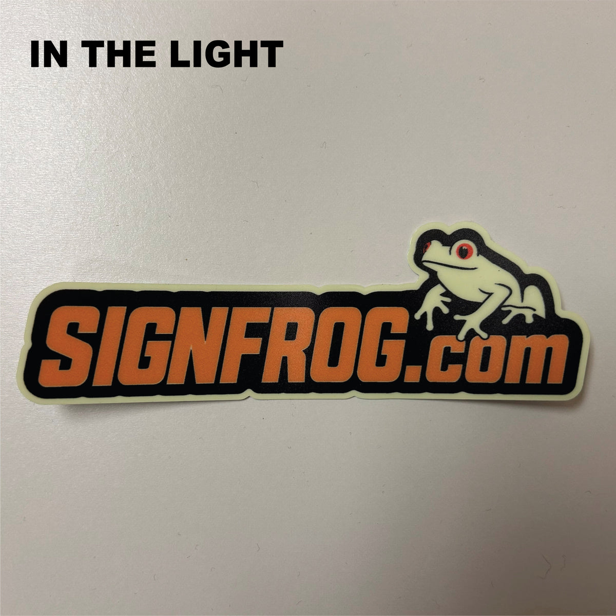A die-cut sticker featuring the text 'SIGNFROG.com' in orange with a black outline, with a cartoon frog on the right and the words 'IN THE LIGHT' printed in black at the top left.