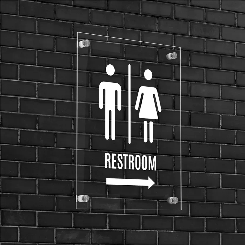 Clear acrylic restroom sign with white icons mounted on black brick wall.
