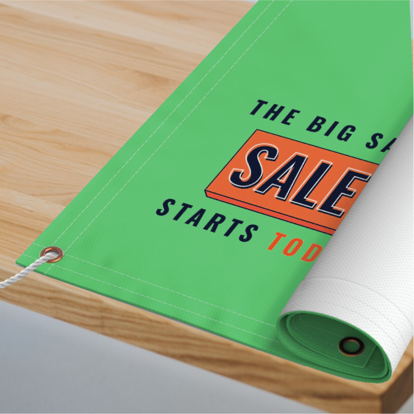 A rolled-up green custom vinyl banner reading The Big Sale Starts Today in bold white and red text, displayed with metal grommets on a wooden table.