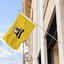 Custom yellow outdoor flag with black logo mounted on a building wall using a silver pole.