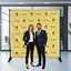 A professional man and woman posing in front of a yellow custom step and repeat logo backdrop banner at a corporate media event.⁠