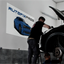 A large custom PVC board sign displaying an automotive logo on a garage wall above a mechanic working on a vehicle.⁠