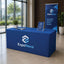 A blue ExpoNova branded table cover on a rectangular table, with a matching vertical banner and acrylic sign in a modern indoor event space with large windows and plants.