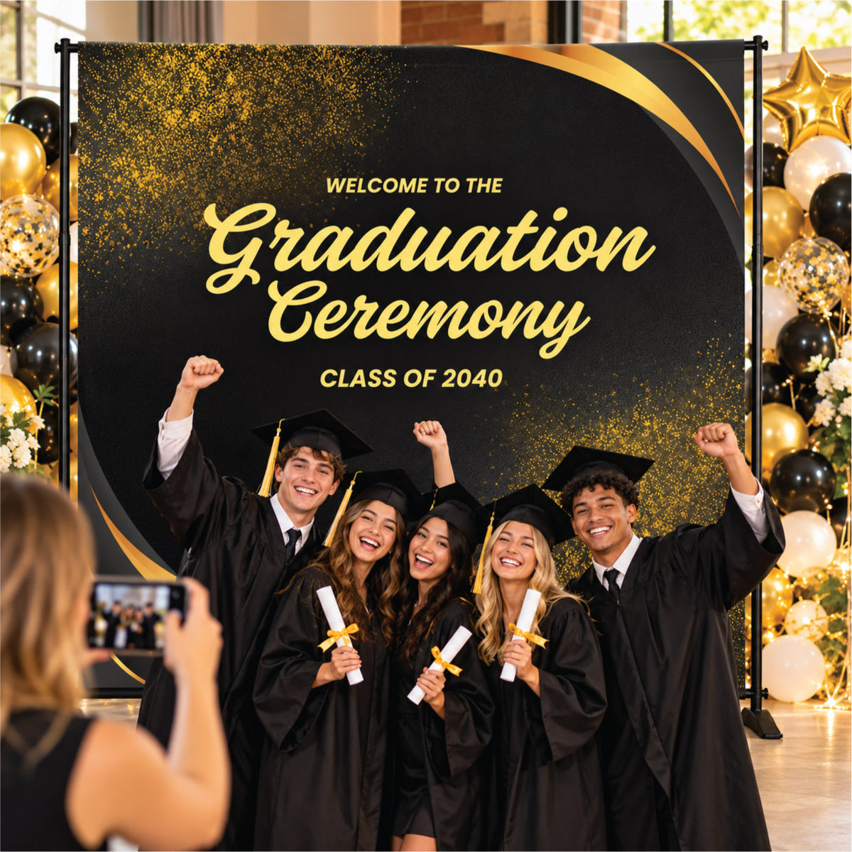 Group of graduating students celebrating at a formal commencement ceremony in front of a black and gold step and repeat backdrop banner.