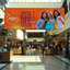 A large horizontal indoor retail promotion vinyl banner hanging from the ceiling over a busy shopping mall corridor.