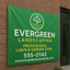 A large green custom vinyl banner advertising landscaping services mounted to an exterior red brick building wall.⁠