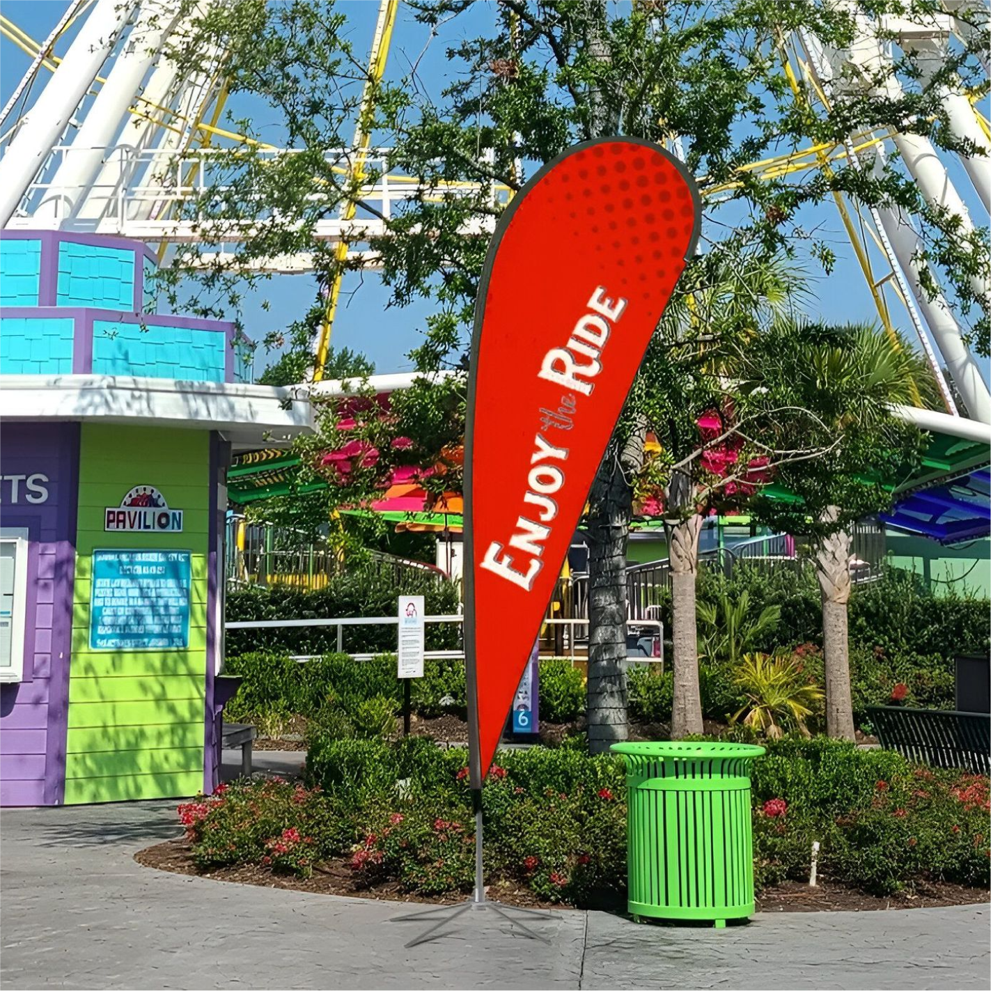 A bright red custom teardrop flag standing outside next to a green trash can with an amusement park ride background.⁠