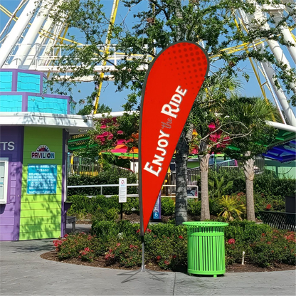 A bright red custom teardrop flag standing outside next to a green trash can with an amusement park ride background.⁠