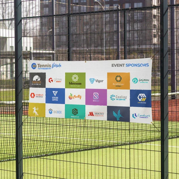 A white multi-logo corporate sponsor vinyl banner attached cleanly to a chain-link sports field fence.⁠