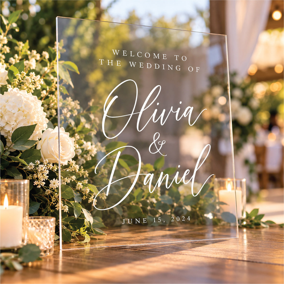 An elegant clear acrylic wedding welcome sign featuring custom white lettering for Olivia and Daniel with a June 15, 2024 wedding date, displayed outdoors surrounded by white floral arrangements and warm candlelight.