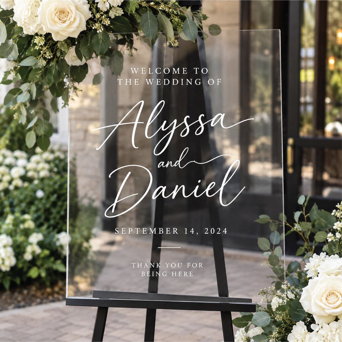 Elegant clear acrylic wedding welcome sign featuring custom white lettering with the couple's names and wedding date, displayed on a black easel outdoors.