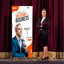 An orange corporate presentation X-banner stand positioned on a stage next to a speaker in front of a red curtain background.⁠
