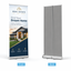 A dual-panel retractable banner stand shown in front and back views on a light gray background. The left panel is printed with a real estate advertisement featuring a house photo and the text 'REAL ESTATE BOY - SELL INSTANTLY.
