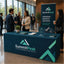 A branded SummitPeak exhibition table and matching vertical banner stand in a modern indoor event space with people conversing in the background. The table is dark teal with large SummitPeak logos and geometric accents, and the banner repeats the brand name and slogan 'Strategy.