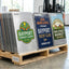 A pallet stacked with custom aluminum novelty signs featuring various business and community logos, including 'Bayport Community Garden' and 'Oak Street Yacht Club', in a warehouse setting.