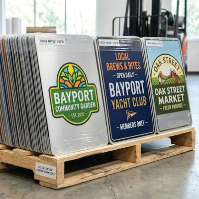A pallet stacked with custom aluminum novelty signs featuring various business and community logos, including 'Bayport Community Garden' and 'Oak Street Yacht Club', in a warehouse setting.